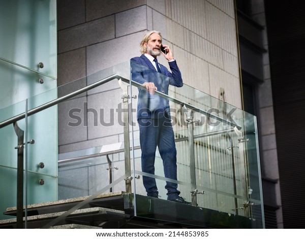 mature\
caucasian corporate business man standing on top of stairs making a\
call using cellphone in modern office\
building