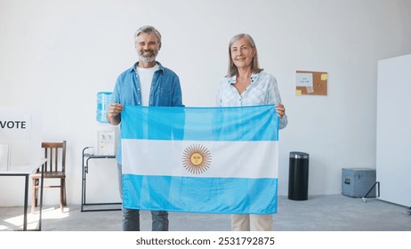 Mature Caucasian couple looking at each other before smiling to camera. Holding large Argentinian flag. Smiling with joy. Motivating others to come vote and complete their duty. Voting location. - Powered by Shutterstock