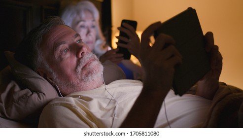 A mature Caucasian couple lying in bed at night engaged in the unhealthy habit of using smart devices at bedtime. - Powered by Shutterstock