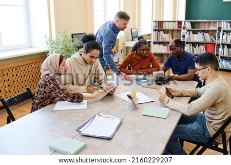 Mature Caucasian English language teacher helping immigrant students with doing task during lesson in library