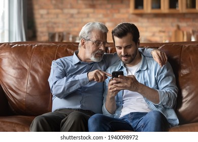 Mature Caucasian father and adult son relax on sofa at home using modern smartphone gadget. Old 60s dad and grownup man kid rest on couch in living room browse internet on cellphone device. - Powered by Shutterstock