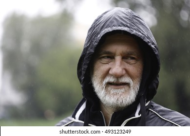 Mature Caucasian Gray Man With Beard And Mustache In A Park Standing In The Rain Wearing Hooded Black Sport Jacket Looking Away From The Camera With Blurry Background