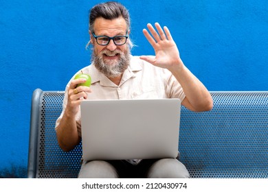 Mature Caucasian Man Eating Apple Waving Hello On A Video Call In Park Bench.