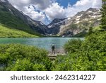 Mature Caucasian man, hiker standing on the edge of a lake in contrast to the water looking at a distant snow capped mountain pass, Gunsight Lake, Gunsight Pass, Glacier NP, Montana