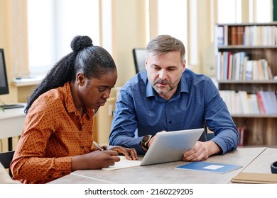 Mature Caucasian teacher sitting next to his Black student explaining her how to do task and showing something on digital tablet - Shutterstock ID 2160229205
