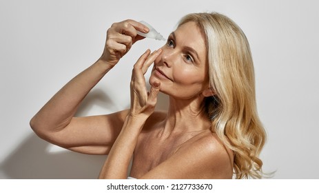 Mature Caucasian Woman Dripping Eye Drops In Her Eye. Cropped Image Of Serious Blonde Female Person Looking At Camera. Health Treatment And Care. Isolated On White Background. Studio Shoot