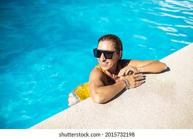 Mature Caucasian Woman Enjoying In Blue Swimming Pool. She Is Wearing Black Sunglasses And Yellow Swimwear