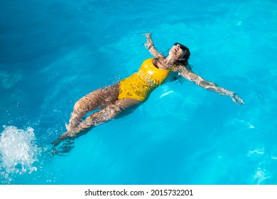 Mature Caucasian Woman Enjoying In Blue Swimming Pool. She Is Wearing Black Sunglasses And Yellow Swimwear