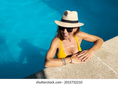 Mature Caucasian Woman Enjoying In Blue Swimming Pool. She Is Wearing Black Sunglasses, Yellow Swimwear And Hat