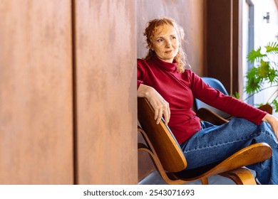 Mature Caucasian woman sitting in modern chair, wearing red sweater and jeans. She appears thoughtful and relaxed, surrounded by warm sunlight and indoor plants. Atmosphere is calm and introspective. - Powered by Shutterstock