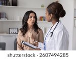 Mature Caucasian woman visiting young African female doctor for consultation, telling about healthcare, sickness symptoms. Physician filling medical form, listening to patient in clinic office