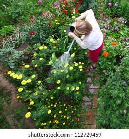 Mature Caucasian Woman Watering Her Green Garden With A Lot Of Flowers