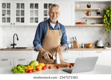 Mature Chef. Smiling Senior Man In Apron Cooking Food In Kitchen, Handsome Elderly Gentleman Using Laptop While Preparing Healthy Vegetable Meal At Home, Grating Zucchini And Looking At Camera - Powered by Shutterstock