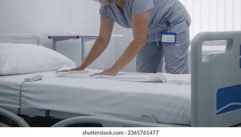 Mature cleaner changes bedclothes and puts clean pillow on bed. Nurse makes beds in hospital ward and prepares it for new patients in bright hospital ward. Medical staff work in modern medical center. - Powered by Shutterstock