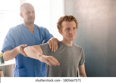 Mature Clinician In Blue Uniform Supporting Bent Arm Of Young Patient During Physiotherapeutic Training In Modern Rehabilitation Clinics