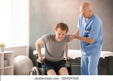 Mature Clinician In Uniform Helping Young Man In Pain To Sit In Wheelchair After Rehabilitation Training While Supporting His Arm And Elbow