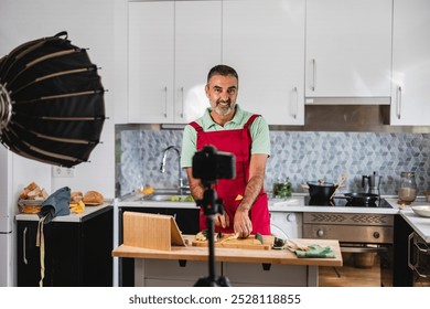 Mature content creator is using a camera on a tripod to film himself preparing food in his kitchen, he's wearing an apron and smiling - Powered by Shutterstock