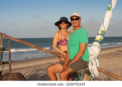 Mature Couple At The Beach In Combuco, Brazil, Sitting On A Jangada A Fishing Boat Typical In Brazil