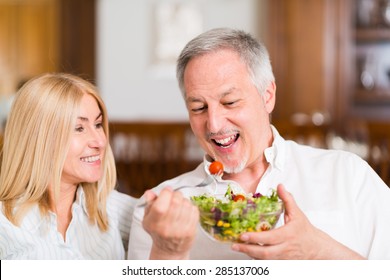 Mature Couple Eating A Salad In The Living Room. Shallow Depth Of Field, Focus On The Man