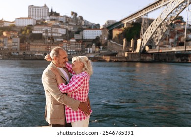 Mature couple embraces by the riverside, smiling at each other with affection. Behind them, a picturesque cityscape and a striking bridge enhance the romantic atmosphere during sunset. - Powered by Shutterstock