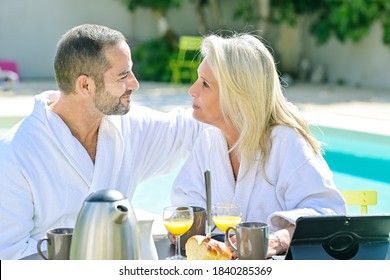 Mature Couple Having Breakfast In Bathrobe Outside In Front Of Their Swimming Pool