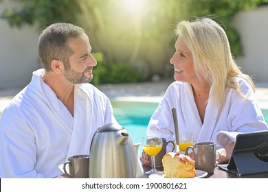 Mature Couple Having Breakfast In Bathrobe Outside In Front Of Their Swimming Pool