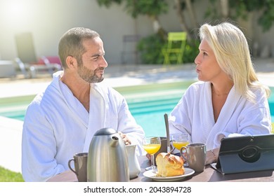 Mature Couple Having Breakfast In Bathrobe Outside In Front Of Their Swimming Pool