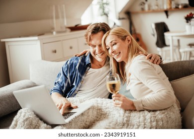 Mature couple having a video call on their laptop while enjoying a glass of wine on the couch in the living room - Powered by Shutterstock