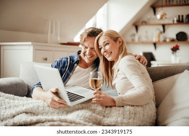 Mature couple having a video call on their laptop while enjoying a glass of wine on the couch in the living room - Powered by Shutterstock