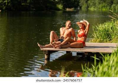 Mature couple joyfully relaxing on a wooden dock, woman adjusting hat and man smiling at her, in a serene lake setting at summer in germany - Powered by Shutterstock