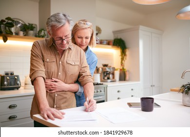 Mature Couple Reviewing And Signing Domestic Finances And Investment Paperwork In Kitchen At Home