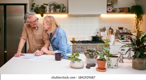 Mature Couple Reviewing And Signing Domestic Finances And Investment Paperwork In Kitchen At Home