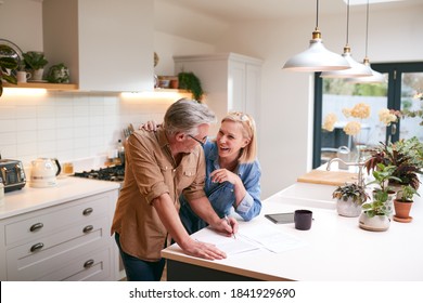 Mature Couple Reviewing And Signing Domestic Finances And Investment Paperwork In Kitchen At Home