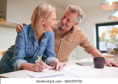 Mature Couple Reviewing And Signing Domestic Finances And Investment Paperwork In Kitchen At Home