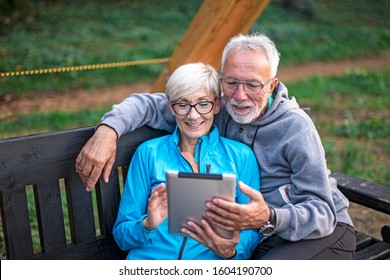 Mature Couple Sitting At Banch In Par And Looking At Tablet And Having Fun