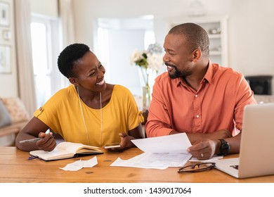 Mature Couple Sitting And Managing Expenses At Home. Happy African Man And Woman Paying Bills And Managing Budget. Black Smiling Couple Checking Accountancy And Bills While Looking At Each Other.