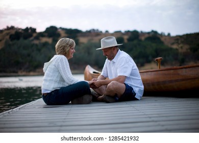 Mature Couple Sitting On Lake Pier Holding Hands Next To A Canoe