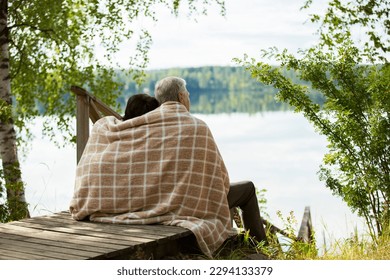 Mature couple sitting on wooden steps and hugging. They are wrapped in blanket, smiling and looking at beautiful view - forest and lake. Happy senior couple embracing each other  - Powered by Shutterstock