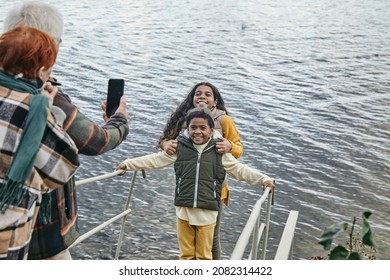Mature couple with smartphone photographing their two cute interracial grandchildren while spending weekend by lake together