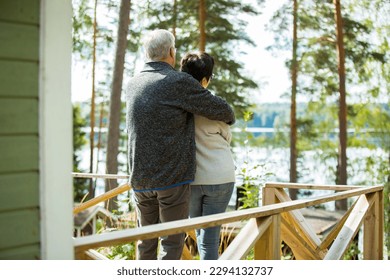 Mature couple standing on porch and hugging. They are smiling and looking at the beautiful view - forest and lake. Happy senior couple embracing each other on the wooden terrace of the house.  - Powered by Shutterstock