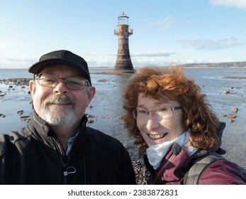 Mature couple take a selfie on the beach with Whiteford Lighthouse behind. Whiteford lighthouse is a wave-swept cast-iron lighthouse and an important work of engineering and 19th century architecture. - Powered by Shutterstock