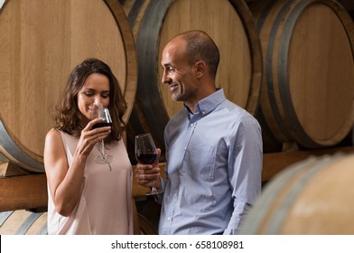 Mature Couple Tasting A Glass Of Red Wine In A Traditional Cellar Surrounded By Wooden Barrels. Happy Mature Woman Smelling A Glass Of Red Wine. Loving Couple Tasting Wines In Winery Cellar.