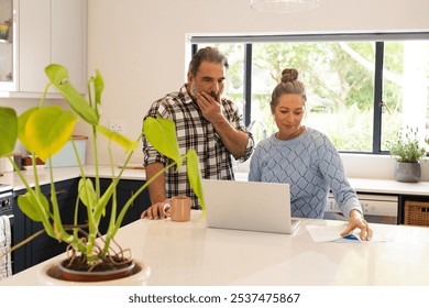 Mature couple using laptop in kitchen, reviewing documents and drinking coffee, at home. Seniors, technology, retirement, lifestyle, finances, home - Powered by Shutterstock