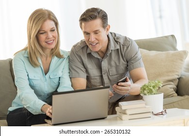 Mature Couple Using Laptop And Mobile Phone While Sitting On Sofa At Home