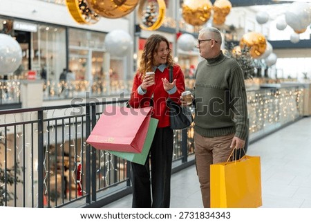 Mature couple walking and talking while holding coffee cups and shopping bags, enjoying a day out in a festive decorated shopping mall during holiday season