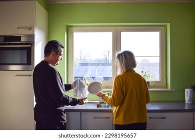 Mature couple washing dishes together in kitchen. Sharing time together. Rinsing and drying plates. People in the morning smiling. Solar panels in the background. Morning routine together. - Powered by Shutterstock