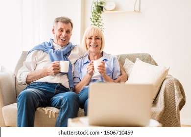 Mature Couple Watching Film On Laptop And Drinking Coffee At Home, Enjoying Weekend Together