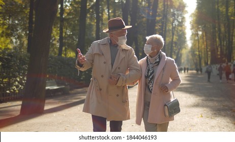 Mature Couple Wearing Sterile Face Masks Walking In Autumn Park. Portrait Of Elegant Aged Man And Woman In Protective Mask Holding Hands Strolling Outdoors. Autumn Vibes