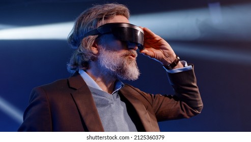 Mature Developer In Augmented Reality Glasses Gesturing And Touching Something In The Air At A Meta Universe Event In Front Of An LED Screen, Explaining To The Audience How Virtual Reality Devices