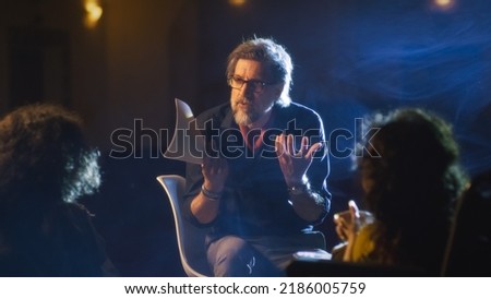 Mature director on a dim theater stage talking to the actor and actress with disability who use electric wheelchair about the performance script, and showing how to act more emotionally